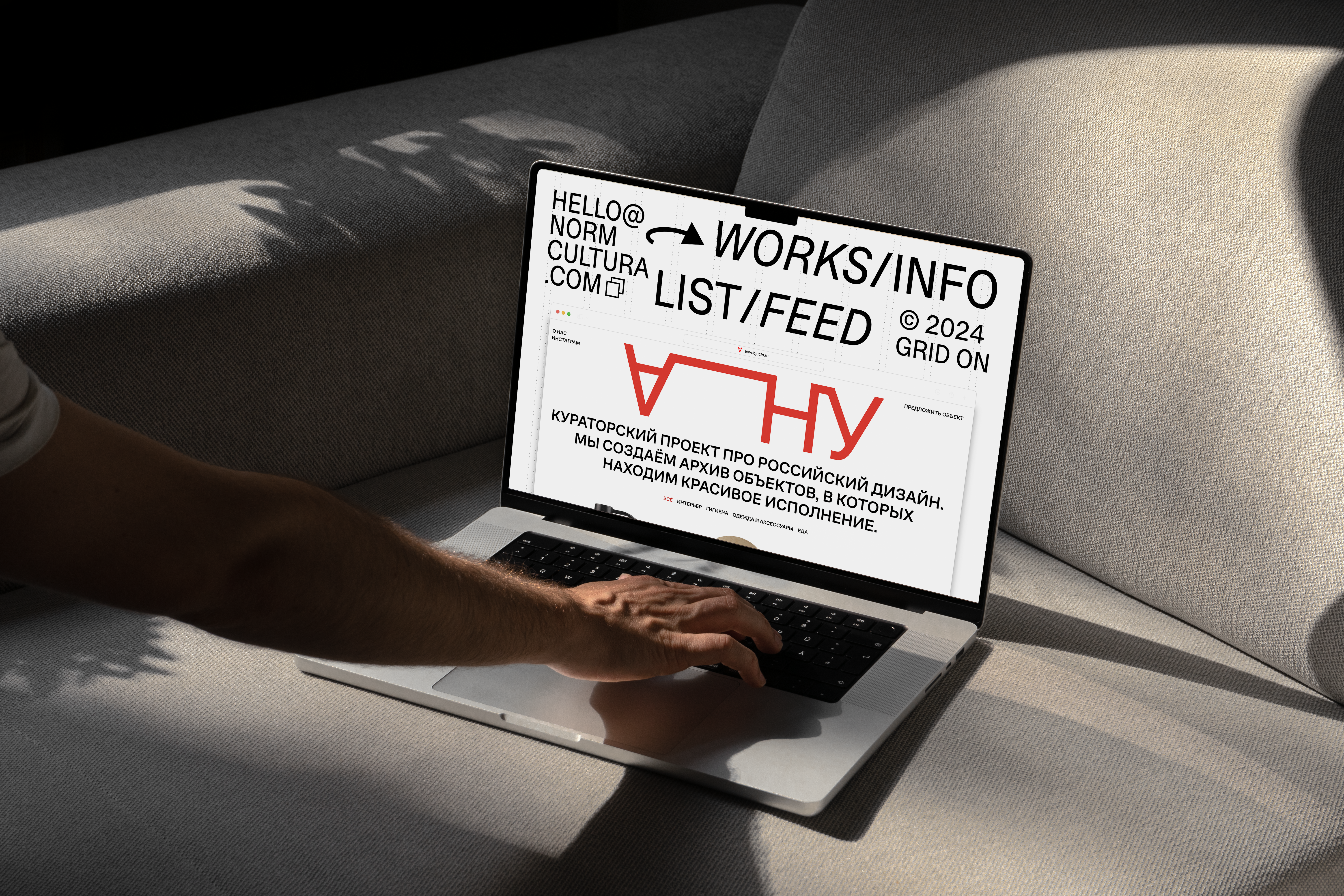 Laptop on gray sofa shows Norm Cultura portfolio homepage with bold “WORKS/INFO LIST/FEED” typography